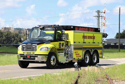 A yellow and black BX tanker drives on an asphalt road in an industrial area with train tracks and three smoke stacks in the background.