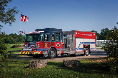 A red and grey Pierce rescue pumper is parked on an angle with blue sky and green wooded surroundings with an American flag in the background. 