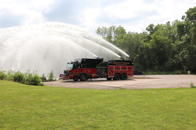 A red and black high flow industrial pumper fire truck sprays water from three nozzles with grass in the foreground and trees in the background. 