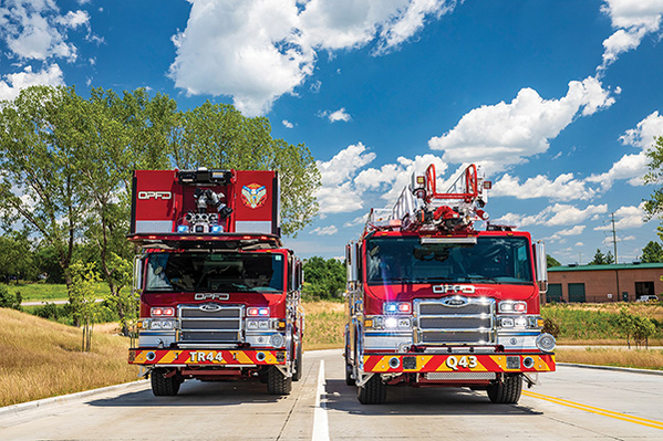 An aerial ladder fire truck and aerial platform fire truck parked next to one another on road on a partly cloudy day