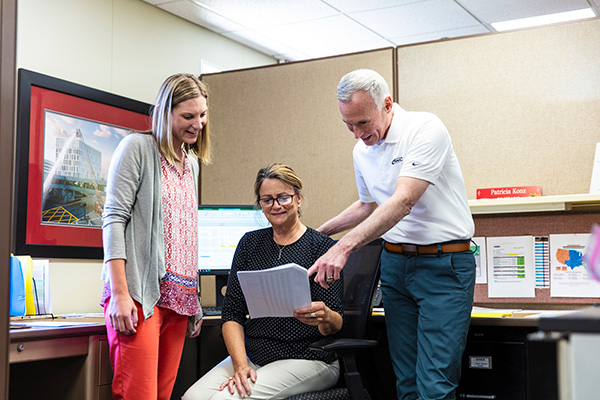 A woman sitting in a chair next to a man and a woman standing in a cubicle looking at printed documents.