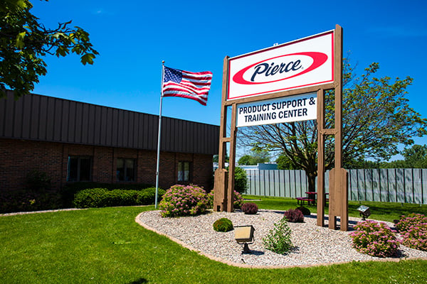 Pierce Product Support and Training Center sign in front of the building next to bushes and an American flag on a sunny day.