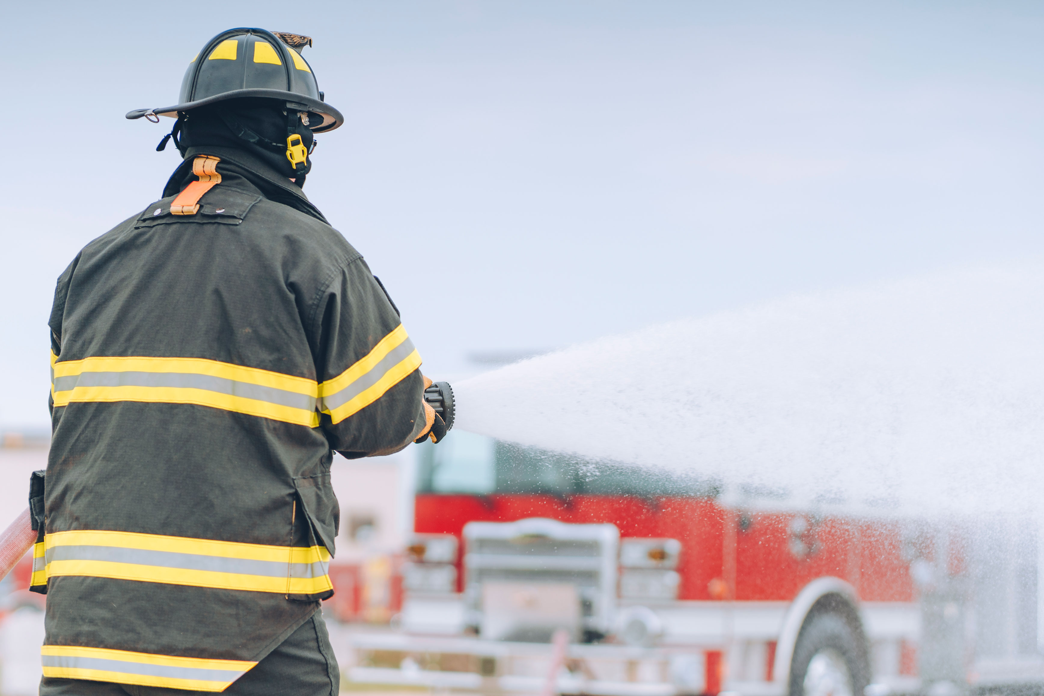 Firefighter standing outside spraying foam from hose nozzle next to a Pierce Fire Truck. 