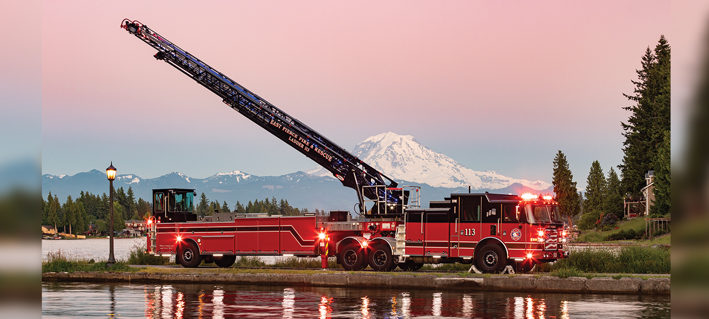 Tiller fire truck parked with emergency lights on and ladder extended against a mountainous background
